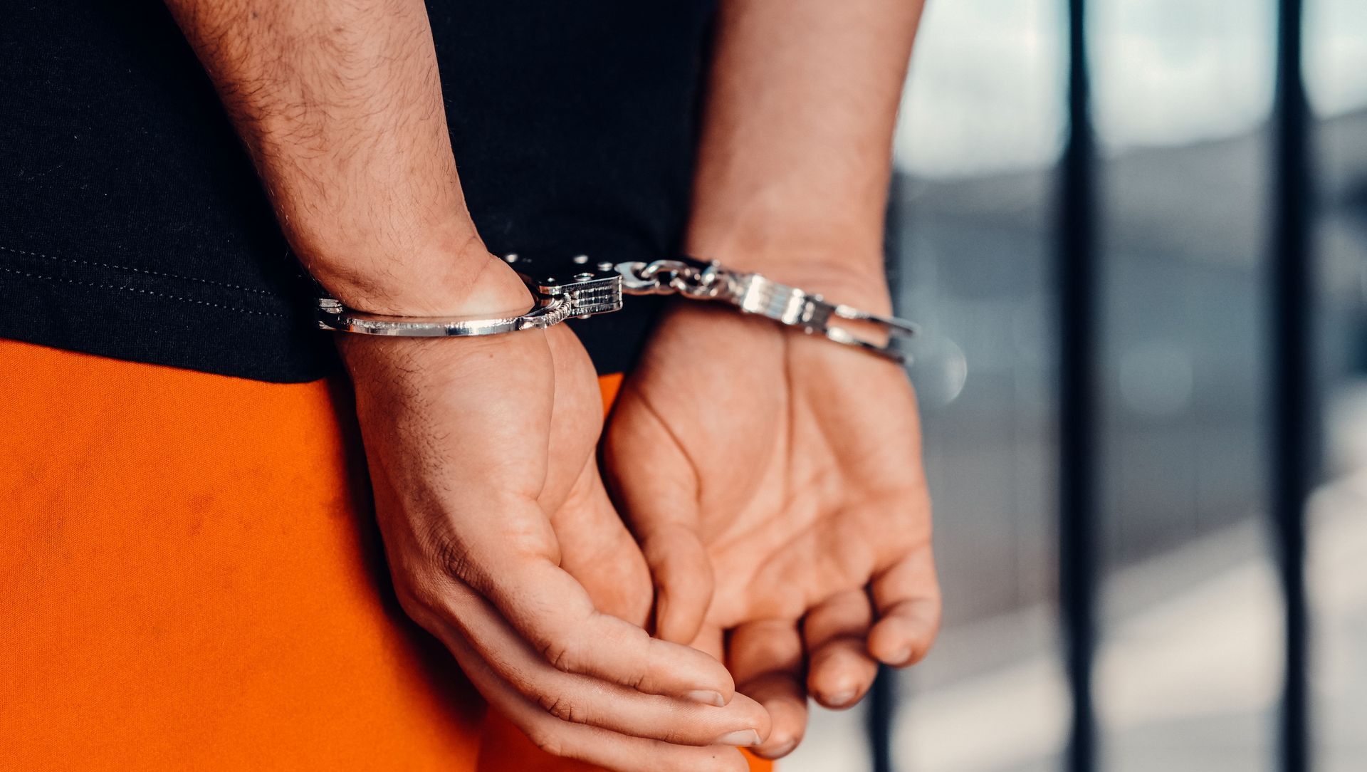 Close-up of person in handcuffs wearing orange attire behind prison bars, symbolizing arrest.